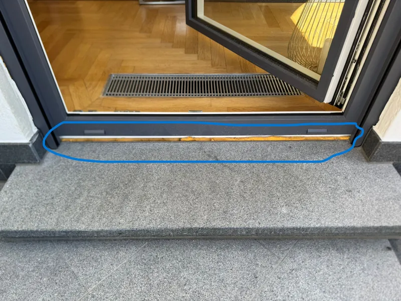 Geöffnete anthrazitfarbene Fenstertür über einer Granitstufe, blau eingekreist: Entlang der Rahmenunterkante liegt die Fuge offen, gelber Schaum ist auf ganzer Breite zu sehen.