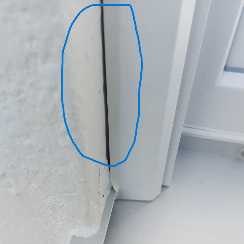 Seitlicher Außenanschluss eines weißen Fensters, blau eingekreist: Zwischen der weißen Leiste am Rahmen und dem Laibungsputz verläuft ein durchgehender dunkler Spalt.