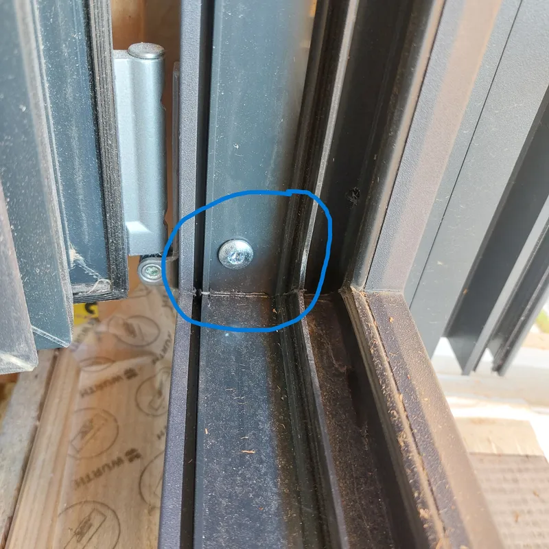 Innenecke eines anthrazitfarbenen Rahmens, blau eingekreist: Die Befestigungsschraube sitzt dicht über der unteren Rahmenecke. Links ein Flügel mit Band.